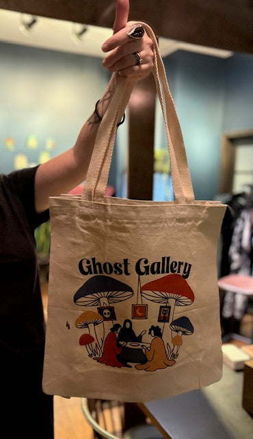 Person holding a tote bag with 'Ghost Gallery' design featuring mushrooms and people around a table.