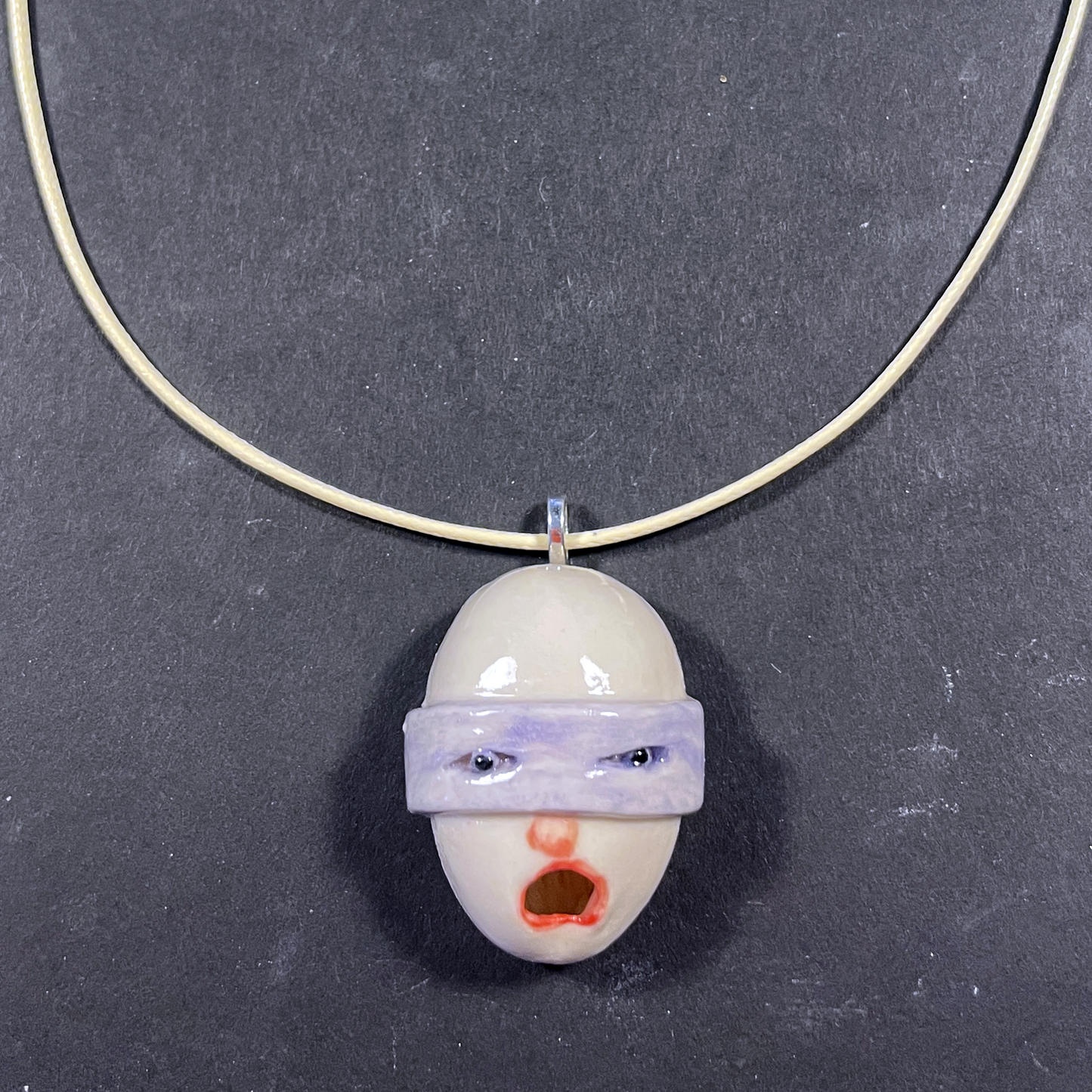 A handmade ceramic pendant necklace featuring a stylized face with a purple blindfold and red mouth, suspended on a thin gold-colored wire chain against a dark gray background.