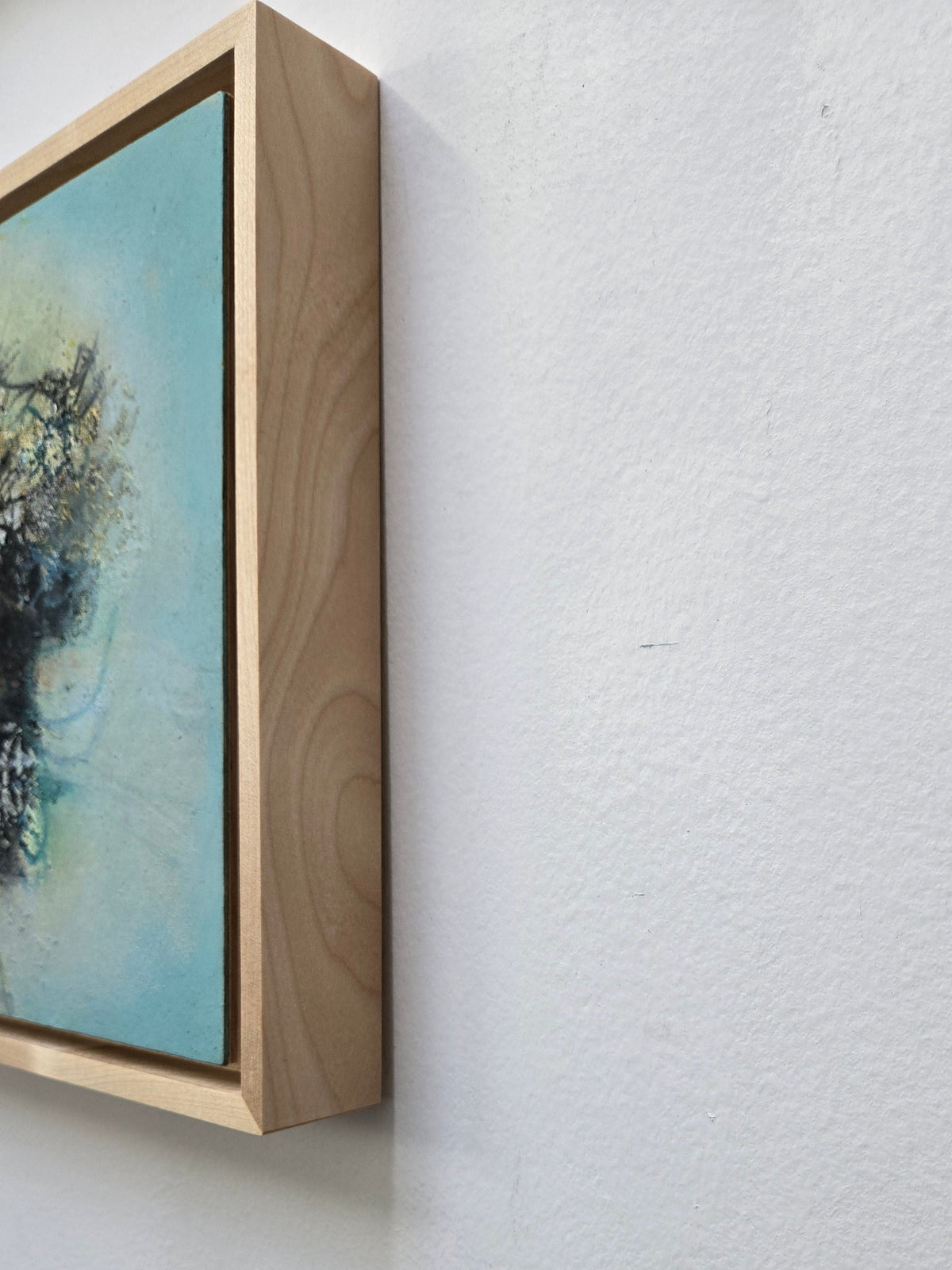 A framed abstract painting with a light blue background and dark tree-like brushstrokes, shown hanging on a white textured wall.