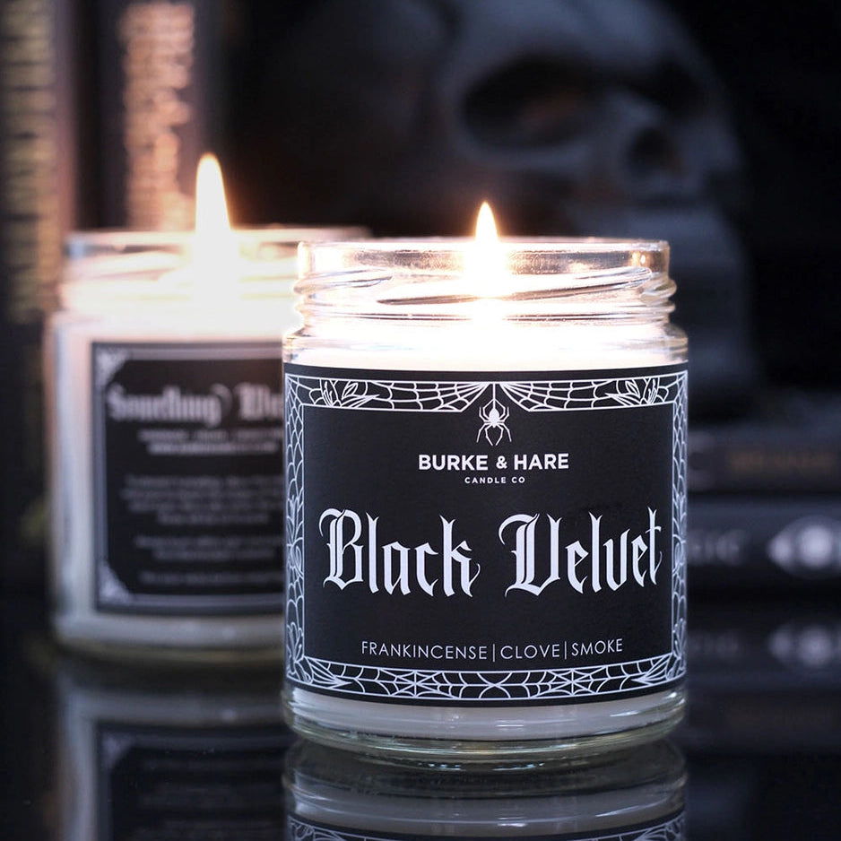 Two lit candles in glass jars labeled 'Black Velvet' with a skull and books in the background.