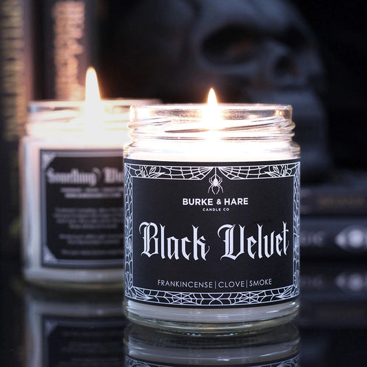 Two lit candles in glass jars labeled 'Black Velvet' with a skull and books in the background.