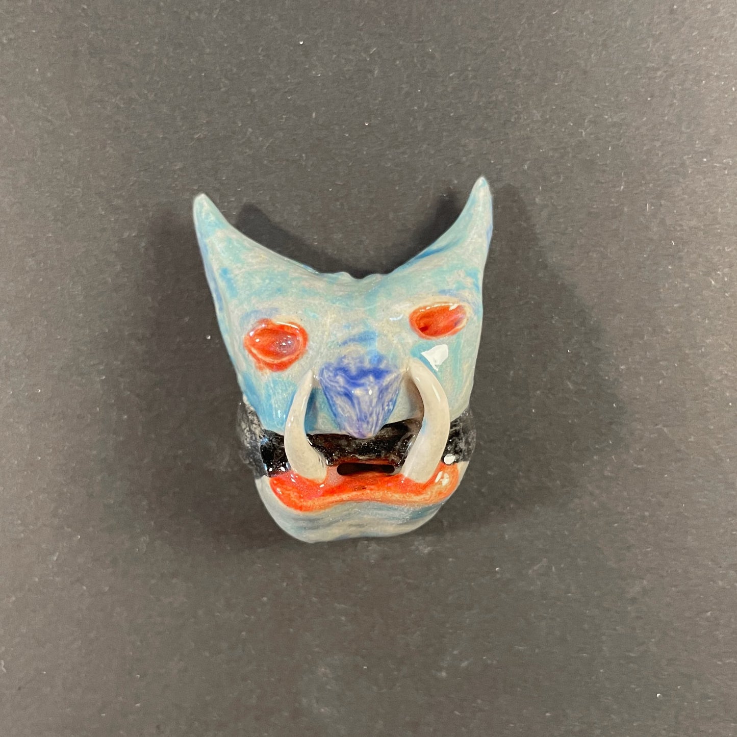 Small ceramic mask with blue face and red accents on a gray background
