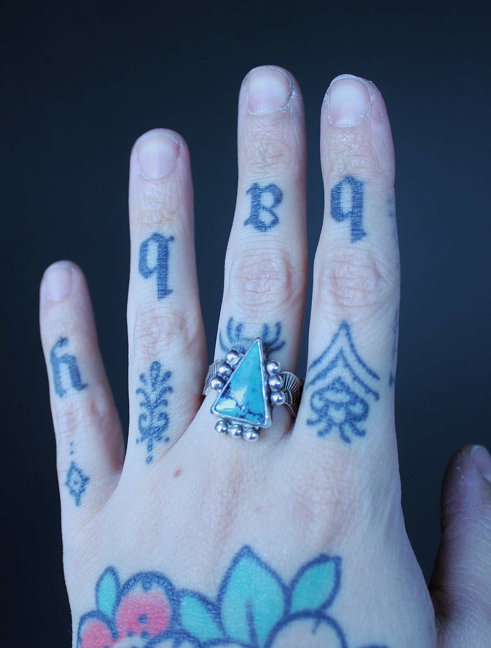 A close-up of a hand with blue tattoos, featuring a silver ring with a triangular turquoise-blue stone in the center, set against a dark background.