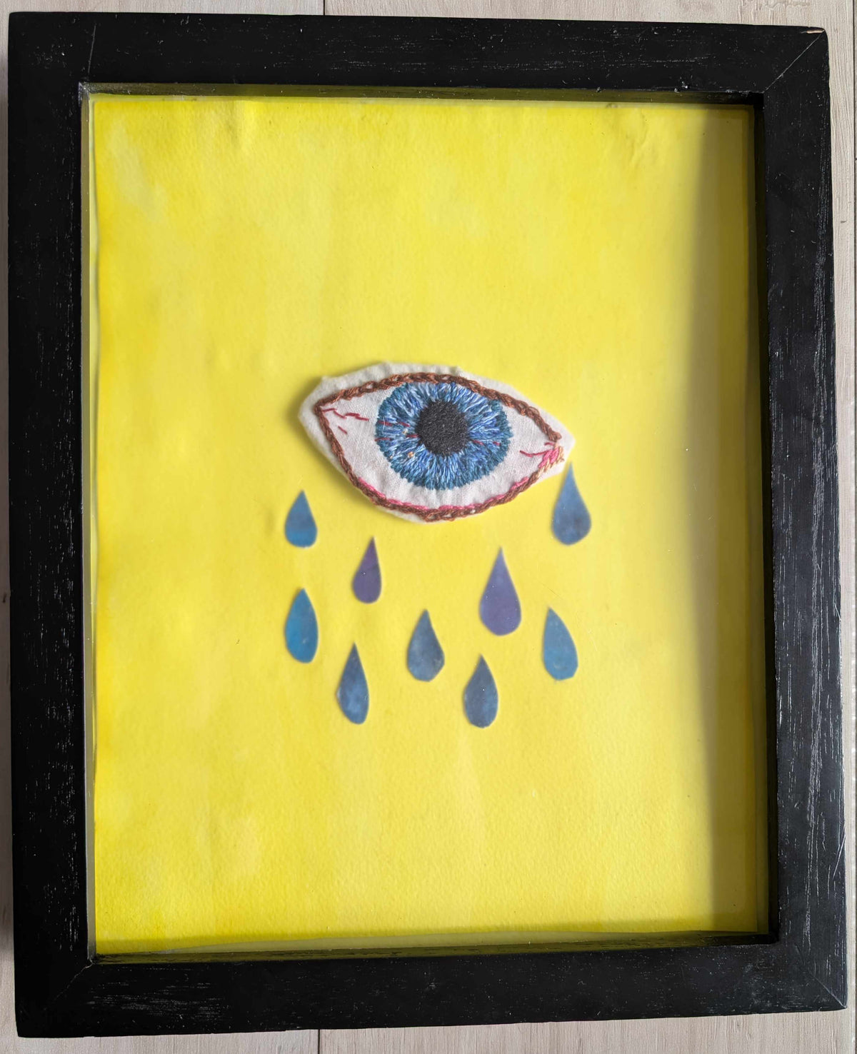 A framed art piece featuring a yellow background with a central embroidered blue eye and blue teardrop shapes, enclosed in a black frame on a light wood floor.