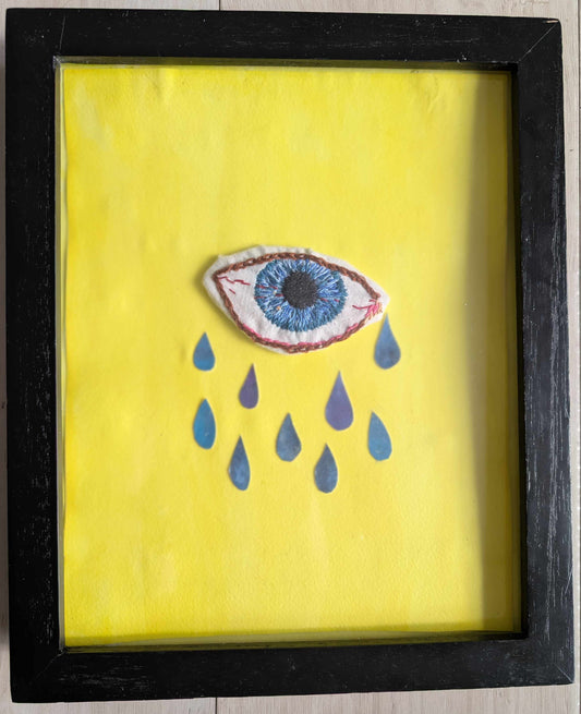 A framed art piece featuring a yellow background with a central embroidered blue eye and blue teardrop shapes, enclosed in a black frame on a light wood floor.
