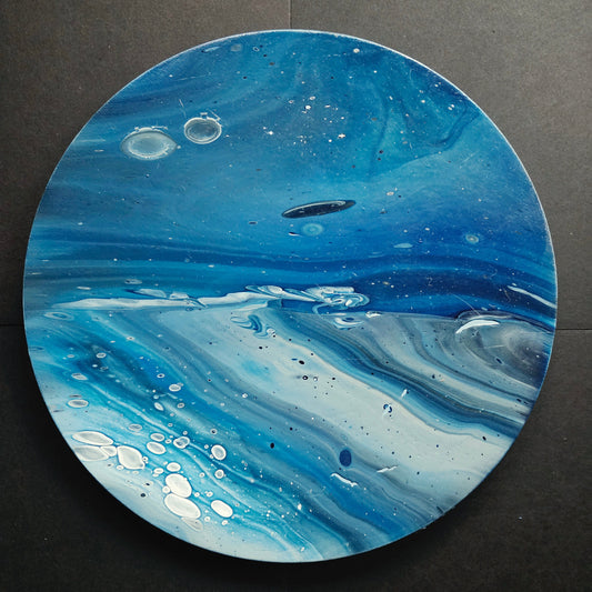 A round abstract fluid art painting featuring swirling patterns of blue, white, and gray with small bubble-like details, displayed on a dark textured background.
