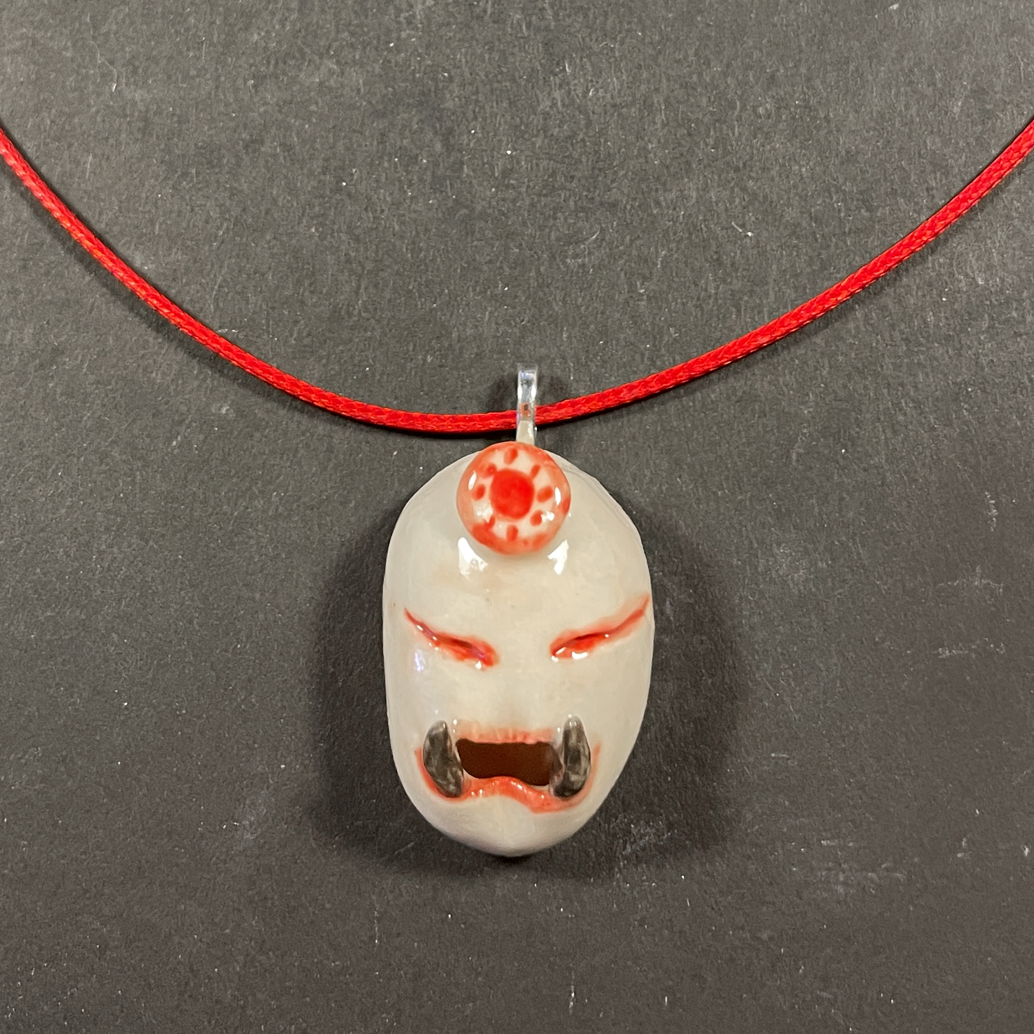 A handmade ceramic pendant on a red cord necklace, displayed on a dark gray textured background.