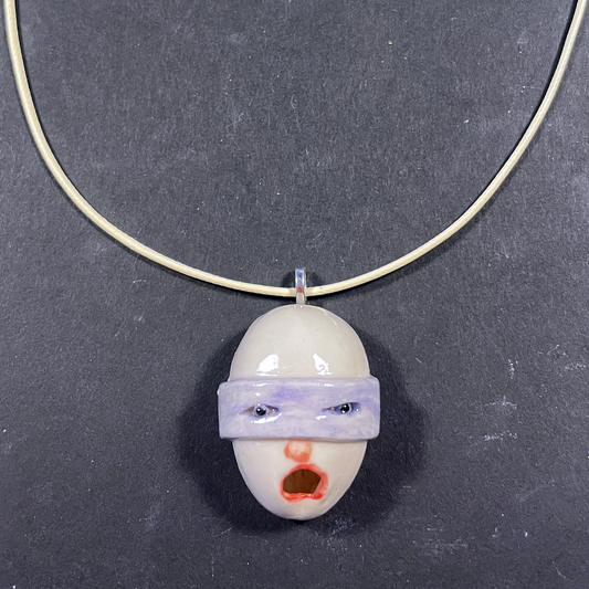 A handmade ceramic pendant necklace featuring a stylized face with a purple blindfold and red mouth, suspended on a thin gold-colored wire chain against a dark gray background.