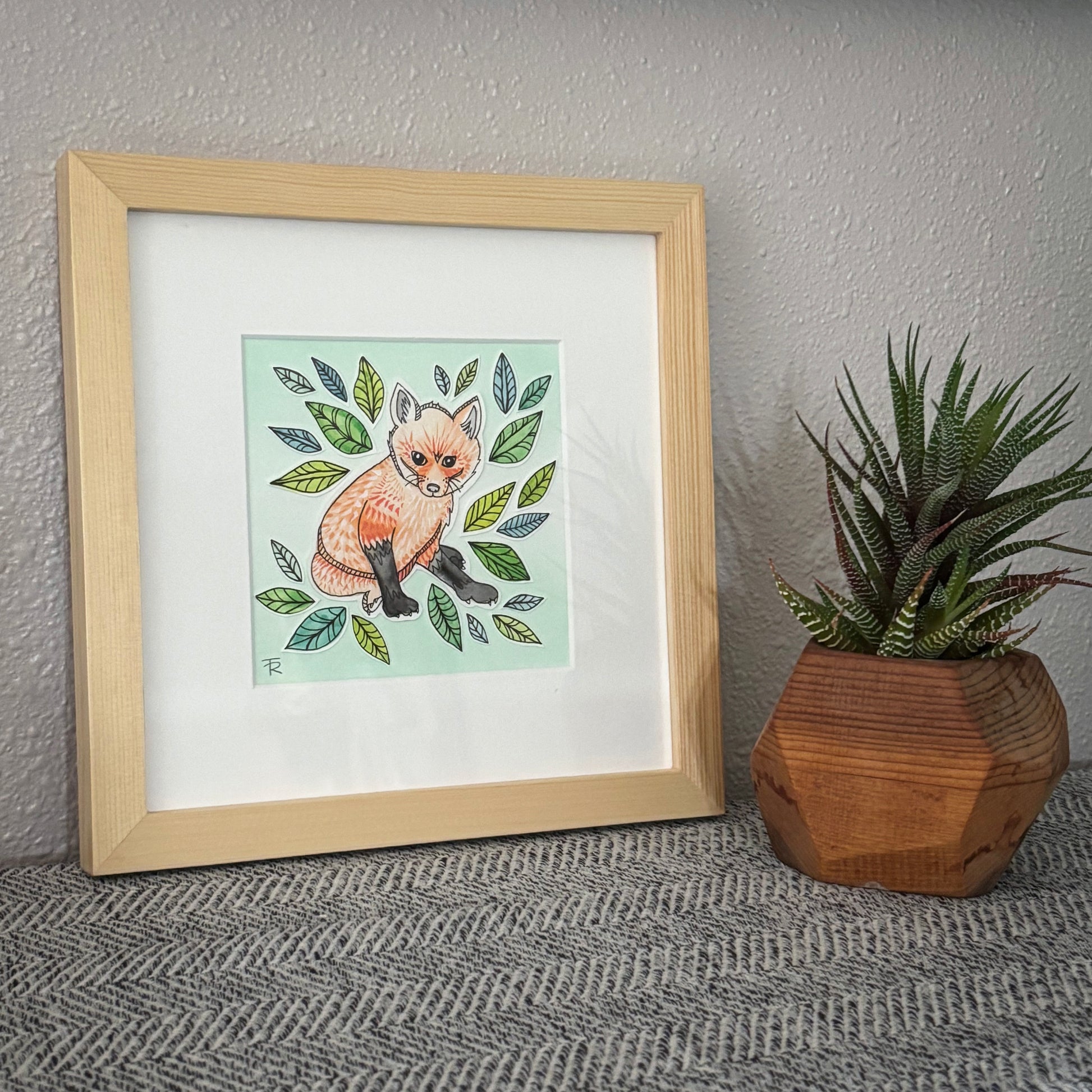 Framed artwork of a fox surrounded by leaves on a textured wall with a plant beside it.