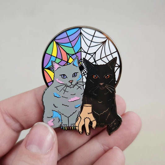 Pin with two cats, one gray and one black, in front of a stained glass and spider web design.