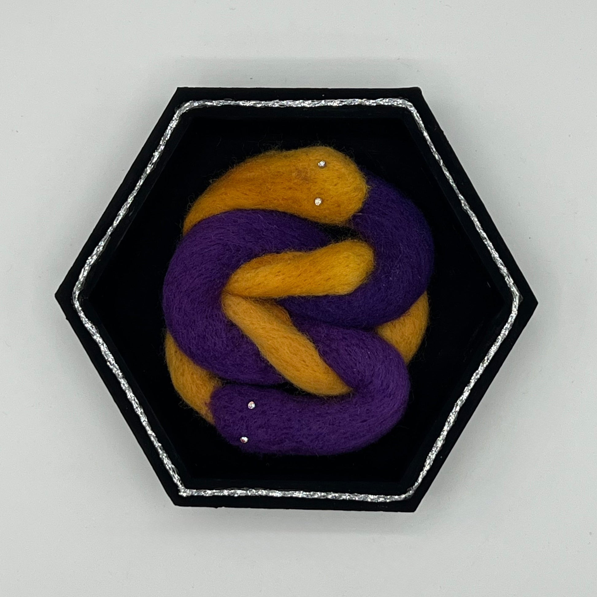 Hexagonal brooch with purple and gold knot design on a light gray background