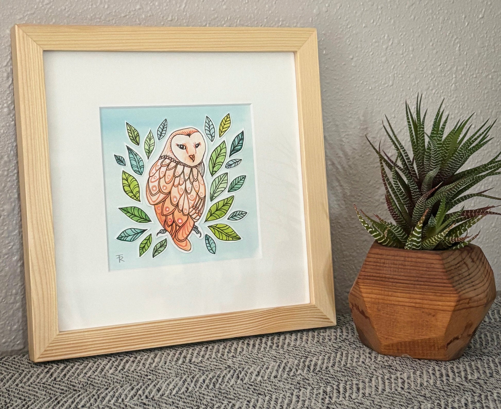 Framed artwork of an owl with leaves next to a small potted plant on a textured surface.