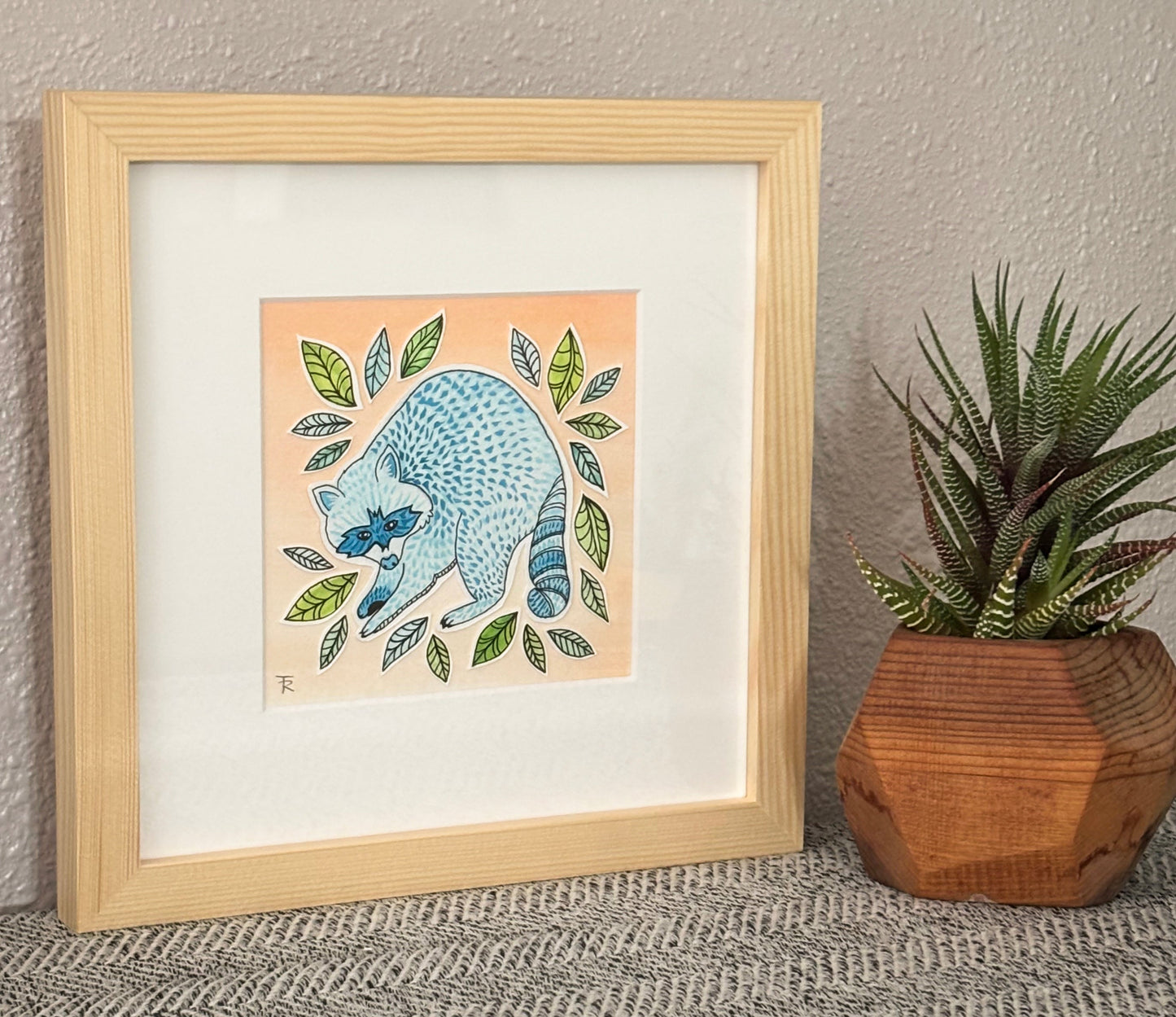 Framed artwork of a blue cat with leaves on a textured wall with a plant.