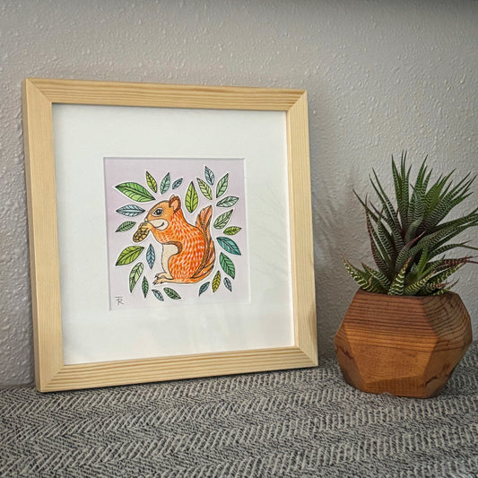 Framed artwork of a squirrel with leaves next to a potted plant on a textured surface.