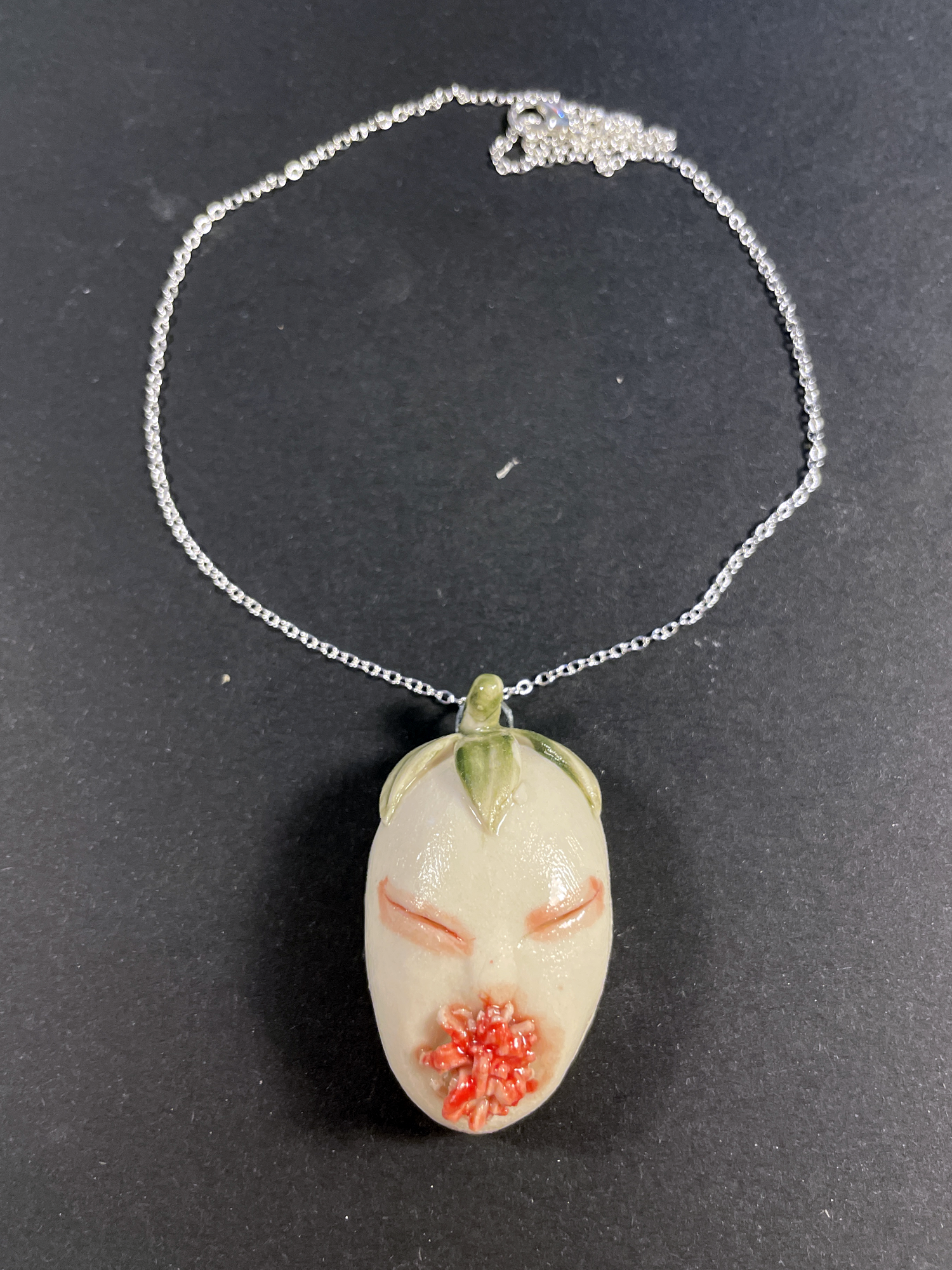 A silver chain necklace featuring a small, hand-painted ceramic or resin pendant shaped like a stylized face with closed eyes, a green leaf detail at the top, and a red flower accent at the bottom, displayed on a dark gray background.